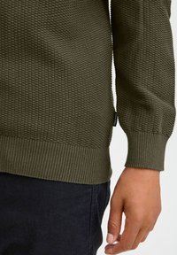 Olivgrüner Strukturpullover mit geripptem Saum und Bündchen, der ein waffelartiges Muster aufweist und ein sichtbares Markenlabel an der Seite hat.