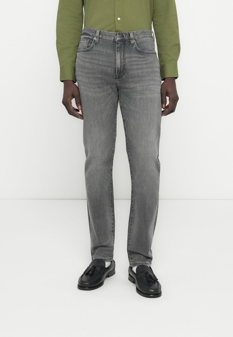 Homme portant un jean slim gris délavé, des mocassins noirs à pampilles et une chemise olive à manches longues boutonnée, debout devant un fond blanc.