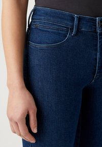 Jeans de denim azul oscuro con corte slim, que presentan un talle alto, bolsillos delanteros y detalles de costura sutiles. Toque de ligero desgaste.