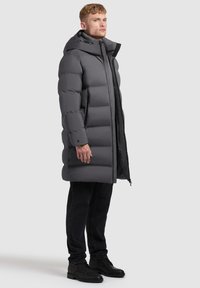 Szary puchowy płaszcz z kapturem, o gładkim wykończeniu, tubularnym kroju i zapinany na zamek z przodu. W zestawie z czarnymi jeansami i butami.