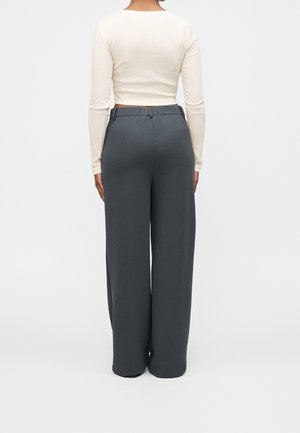 Personne portant un pantalon large gris foncé taille haute et un haut crème ajusté à manches longues, debout de dos à la caméra.