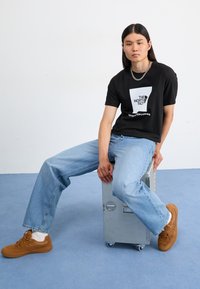 Camiseta gráfica negra con logo blanco, jeans de mezclilla azul claros y zapatillas de ante marrones. Persona sentada sobre una maleta metálica.