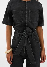 Čierny deninový overal s krátkymi rukávmi, dvoma náprsními vreckami, predným zipsom a viazaným pásom. Má hladkú textúru a jemné švy.