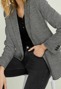 Blazer grigio a quadri con rever strutturati, realizzato in tessuto strutturato. Abbinato a una maglia nera e jeans scuri, con dettagli a bottoni.