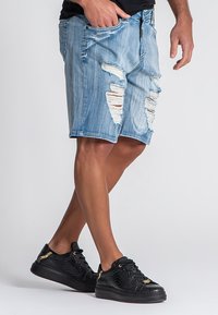 Shorts de mezclilla en azul claro con parches desgastados y bordes desgastados. Combinados con zapatillas negras texturizadas con detalles en dorado.