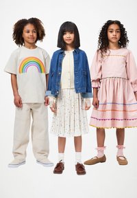 Troje dzieci prezentuje stroje: beżowa koszulka z tęczą, niebieska dżinsowa kurtka na białej plisowanej spódnicy w kolorowe kropki oraz różowa sukienka.