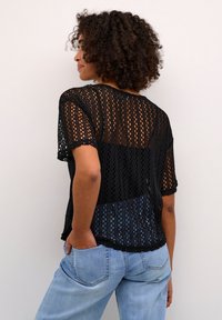 Schwarzes, offenen Strickoberteil mit kurzen Ärmeln und gewelltem Saum, getragen über hellblauen Jeans, was eine lockere Passform und strukturiertes Design zeigt.