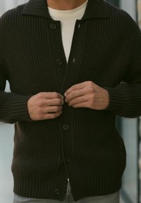 Cardigan en tricot côtelé marron foncé avec col châle, cinq boutons sur le devant, et tissu texturé. Porté par-dessus une chemise blanche.