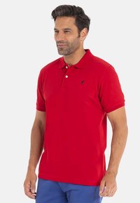 Homme portant un polo rouge à manches courtes avec deux boutons et un pantalon bleu, se tenant contre un fond clair et uni.
