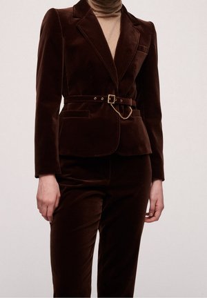 Vrouw die een donkerbruine, fluwelen, getailleerde blazer draagt met bijpassende broek en een dunne riem met een klein goudkleurig kettingdetail.