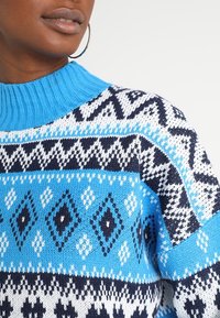 Blauer und weißer gemusterter Strickpullover mit ribbeltem Kragen, der Rautenformen, Zickzack-Muster und kontrastierende marineblaue Akzente aufweist.
