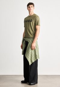 Olivovo zelené bavlnené tričko s odvážnym čiernym logom "HUGO", skombinované s viazanou zelenou kockovanou košeľou a čiernymi širokými nohavicami, doplnené čiernymi čižmami.