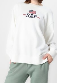 Sweatshirt blanc en tissu doux avec un logo "GAP" brodé en marine et un drapeau américain au-dessus. Silhouette décontractée.