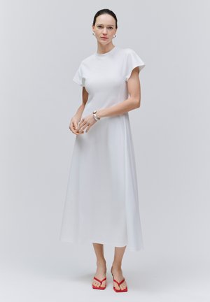 Kobieta stojąca na jednolitym tle, ubrana w białą midi sukienkę z krótkim rękawem i czerwone klapki, z włosami spiętymi do tyłu i neutralnym wyrazem twarzy.