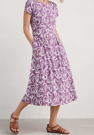 Kvinde iført lilla midi-kjole med hvide abstrakte mønstre, korte ærmer, sidelommer og brune flade sandaler med stropper, stående på en grå gulv.