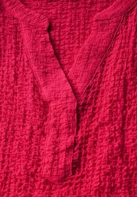 Tejido texturizado en color carmesí con detalle de cuello y tapeta. La superficie presenta un patrón arrugado, añadiendo profundidad e interés visual.