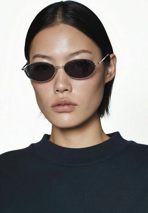 Gros plan d'une personne aux cheveux noirs courts portant des lunettes de soleil ovales foncées et un haut à col rond sombre sur un fond clair uni.