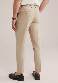 Hombre vestido con pantalones beige a medida, reloj de cuero marrón y mocasines de cuero marrón oscuro, de pie con una mano relajada a un lado.