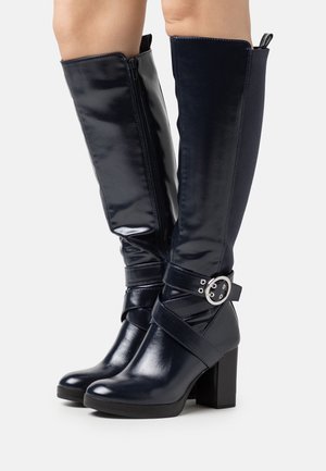 High-heeled, knee-high boots in glossy navy with a side zipper, strap accent, and circular hardware detail on the ankle. Smooth texture.