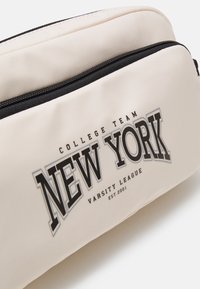 Bolsa de cosméticos de cor creme com textura suave, apresentando um fecho zip preto e uma impressão proeminente de "NEW YORK" em letras negritas.