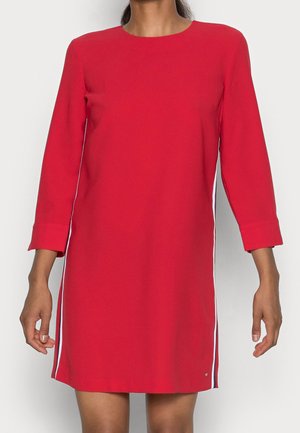 Woman wearing a red, knee-length dress with 3/4 sleeves and white side stripes, standing against a plain light background.