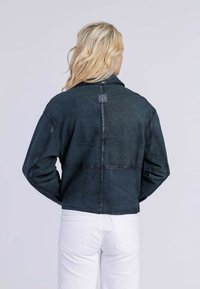 Blonde persoon in een zwart leren jasje en witte broek, staand met de rug naar de camera tegen een effen lichte achtergrond.