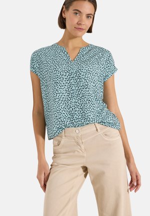Femme portant un chemisier bleu clair à manches courtes avec des points noirs irréguliers et un pantalon beige taille haute, debout devant un fond blanc.