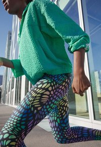 Maglietta verde a maniche lunghe con texture abbinata a leggings neri caratterizzati da un motivo a pois colorato. L'outfit è progettato per un movimento attivo.