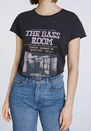 Person iført sort T-shirt med teksten "The Bass Room" og begivenhedsdetaljer, kombineret med højtaljede blå jeans mod en ensfarvet baggrund.
