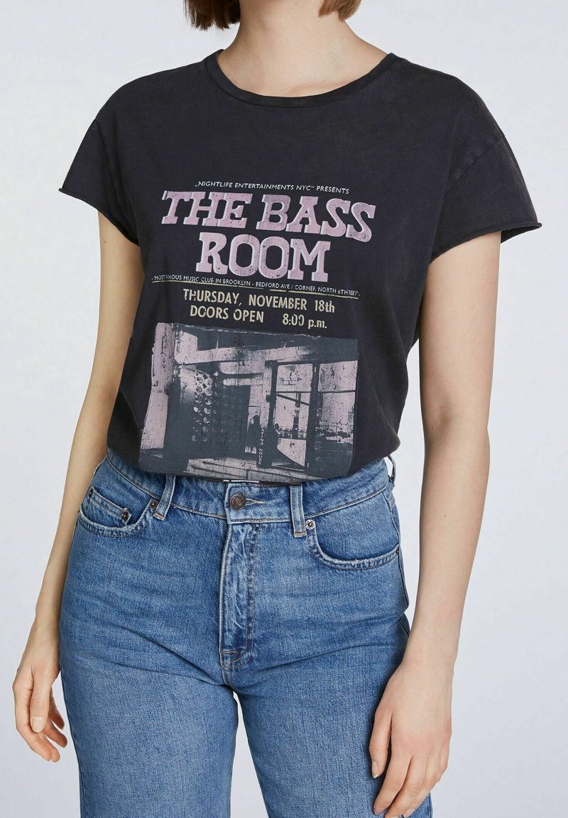 Personne portant un t-shirt noir avec l'inscription "The Bass Room" et les détails de l'événement, associée à un jean bleu taille haute sur un fond uni.