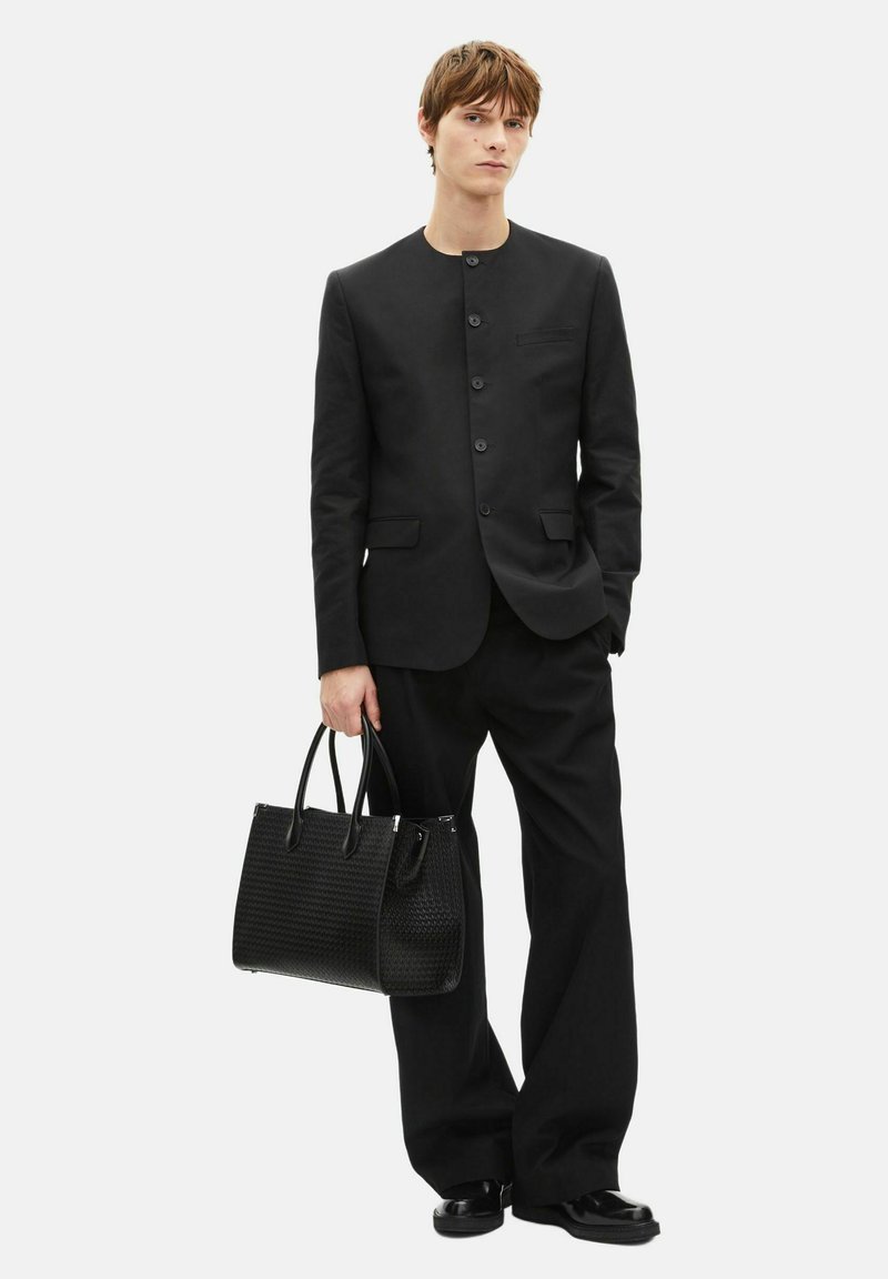 Black tailored jacket with a round neckline, single-breasted buttons, and two pockets. Pair of loose black trousers and woven handbag.