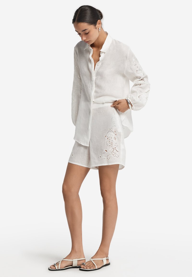 Vit linneskjorta med långa ärmar, knäppning framtill och broderade detaljer. Kombinerad med vita shorts med blommigt broderi och platta sandaler.