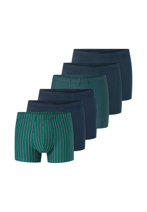 Giovane uomo senza maglietta con pelle chiara e capelli castani che indossa boxer verdi e neri a righe verticali.