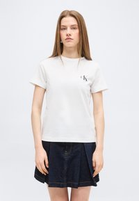 Νεαρή γυναίκα με ίσια μαλλιά που φορεί λευκή μπλούζα Calvin Klein και σκούρα μπλε πλισέ τζιν φούστα, στέκεται μπροστά σε απλό φόντο.