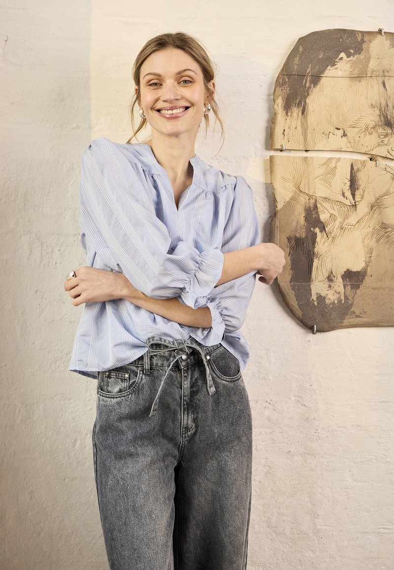Femme souriante en blouse rayée bleu clair et jean gris taille haute, debout les bras croisés devant un mur texturé et une œuvre d'art abstraite en bois.