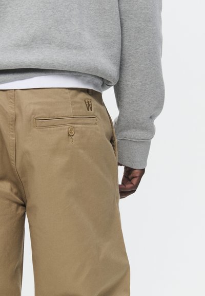 Pantaloni khaki con dettagli a bottoni e tasche posteriori, indossati con una felpa grigia. Presenta una discreta 'W' ricamata vicino alla tasca.