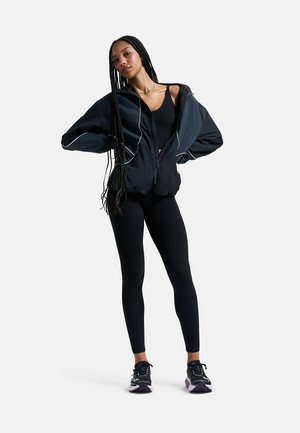 Woman with long braids stands with hands on hips wearing black athletic leggings, black sports bra, black zip jacket, and black sneakers.