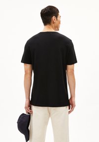 Schwarzes Baumwoll-T-Shirt mit kurzen Ärmeln und Rundhalsausschnitt, von hinten gezeigt. Das Model hält eine marineblaue Kappe und trägt dazu hellbeige Hosen.