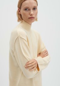 Gestrickter Pullover in Cremefarbe mit hohem, geripptem Stehkragen, lockerem Schnitt und gerippten Details an Bündchen und Saum. Weicher, strukturierter Stoff.