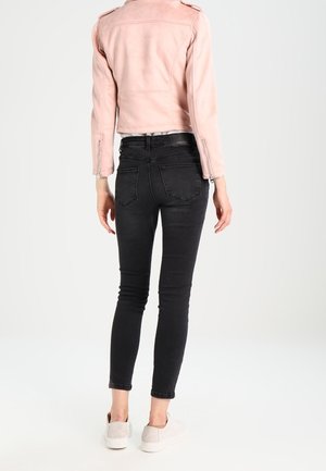Vue de dos d’un mannequin portant une veste rose pâle, un jean slim noir et des baskets blanches sur un fond uni.
