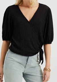 Czarna bluzka zapinana na węzeł z fakturowanej tkaniny, z głębokim dekoltem w serek i bufkami, przewiązana w talii. W zestawie z jasnoniebieskimi dżinsami i złotą bransoletką.