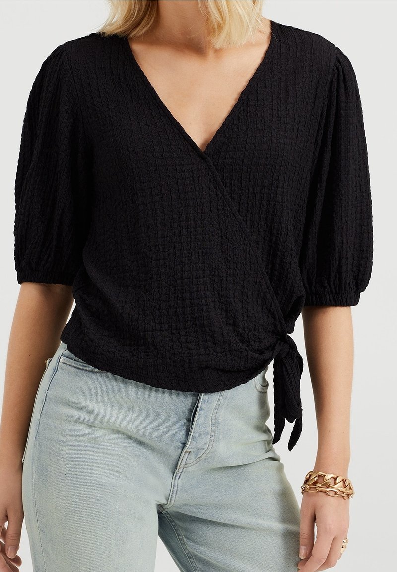 Czarna bluzka zapinana na węzeł z fakturowanej tkaniny, z głębokim dekoltem w serek i bufkami, przewiązana w talii. W zestawie z jasnoniebieskimi dżinsami i złotą bransoletką.