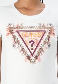 Balta trumpa rankovėmis marškinėliai, kuriuose yra trikampio grafika su gėlių akcentais, auksiniais studais ir "GUESS" logo geltonoje ir šviesiai raudonoje spalvose.
