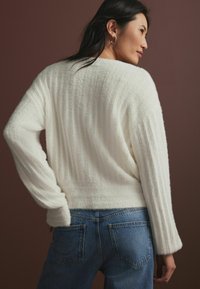 Vit texturerad tröja med ribbad design, som har en avslappnad passform och långa ärmar, kombinerad med högmidjade blå jeans mot en brun bakgrund.