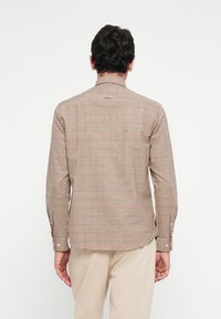 Man facing away wearing a long-sleeve, small checkered shirt in brown and cream with beige pants against a plain white background.