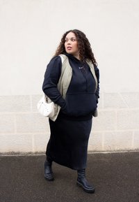 Čierna mikina pod olivovo zeleným vestom, v kombinácii s čiernou maxi sukňou a čiernymi členkovými čižmami. Biely kabelka s kovovými akcentmi. Kučeravé vlasy.