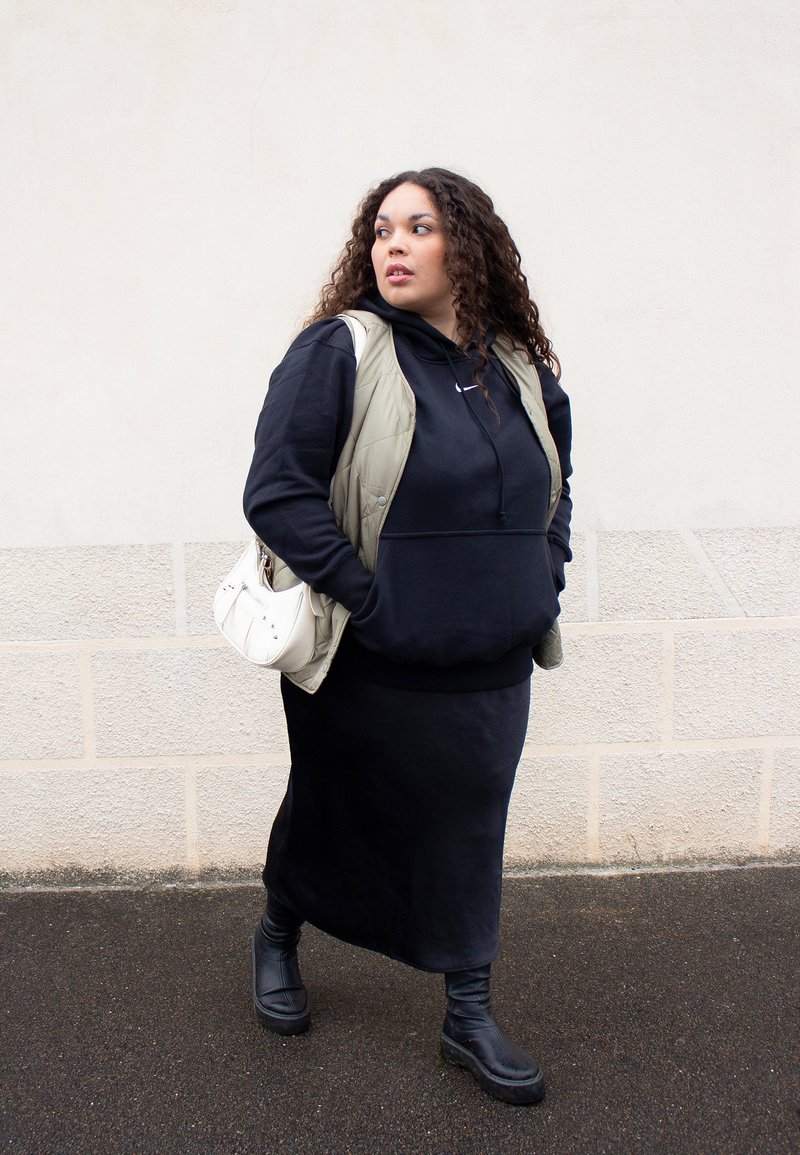 Čierna mikina pod olivovo zeleným vestom, v kombinácii s čiernou maxi sukňou a čiernymi členkovými čižmami. Biely kabelka s kovovými akcentmi. Kučeravé vlasy.