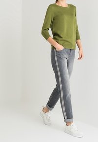 Grüner Strickpullover mit Dreiviertelärmeln, kombiniert mit grauen Jeans mit seitlichen Streifen. Das Modell trägt weiße Sneaker.