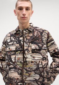 Camo-patterned jacket with a blend of browns and greens, featuring two chest pockets, button closures, and a soft, textured fabric.
