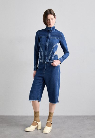Jeansjakke med høy krage og frynser langs kantene, kombinert med knelang denimshorts. Accentueres med lysegule klosshæler og brune sokker.